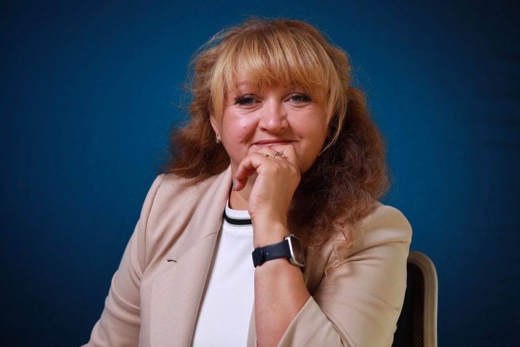 Женщина с улыбкой подпирает подбородок рукой, в бежевом жакете и смарт-часах на синем фоне